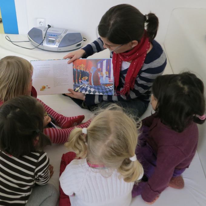 Das Bild zeigt eine Frau. doe vier Kindergartenkindern ein Bilderbuch vorliest.