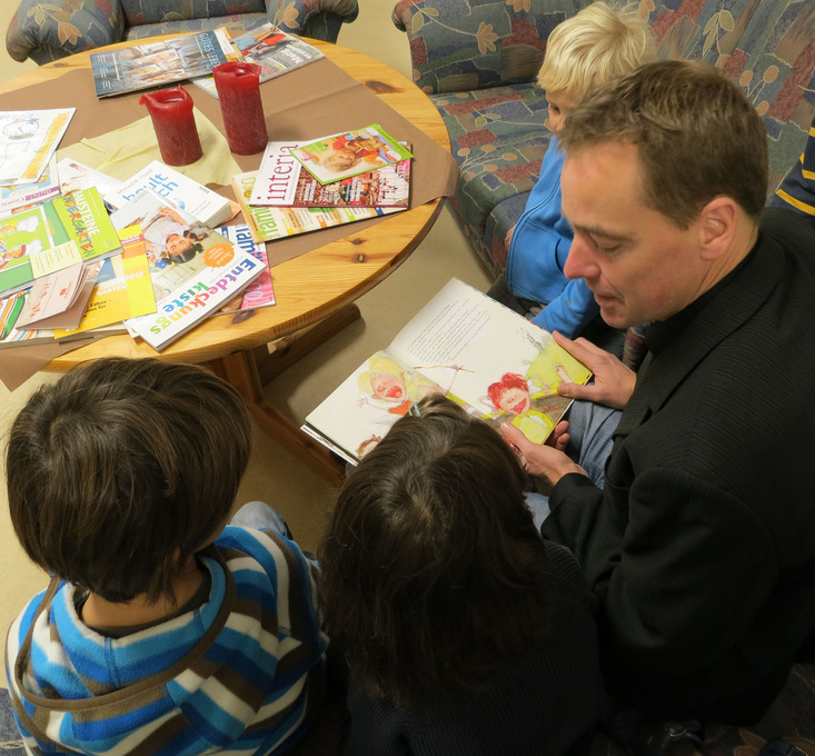 Hier sieht man einem Mann mit einem buch und drei Kindergartenkinder, die zusammen auf einem Sofa sitzen.