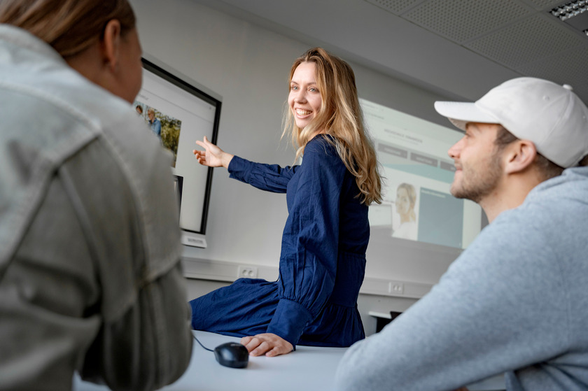 Drei Studierende sitzen zusammen in einem Vorlesungsraum und eine Studentin zeigt etwas auf dem Whiteboard.