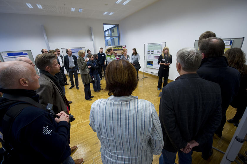 Auf dem Bild sieht man viele Menschen in einem Kreis stehen. In der Mitte steht eine junge Frau vor einem Poster und erzählt etwas.