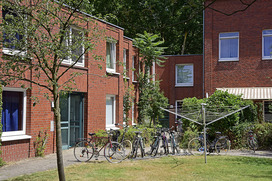 Ein kleiner Innenhof, der von roten Backsteingebäuden umgeben ist. Mehrere Fahrräder sind an einem Fahrradständer abgestellt, der sich neben einem Baum befindet. Eine grüne Wäscheleine steht ebenfalls im Hof. Die Gebäude haben mehrere Fenster, die teilweise mit Pflanzen geschmückt sind. Der Hof wirkt gepflegt und ruhig, und die Umgebung ist von viel Grün umgeben, was eine wohnliche und einladende Atmosphäre schafft.