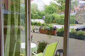 Eine Aussicht von einem Fenster auf einen kleinen Balkon, der mit Pflanzen und Blumen geschmückt ist. Auf dem Balkon steht ein Tisch mit einer grünen Tischdecke und ein grüner Gartenstuhl. Mehrere Blumentöpfe mit unterschiedlichen Pflanzenarten sind auf dem Geländer arrangiert, darunter auch bunte Blumen. Im Hintergrund sieht man eine ruhige Wohngegend mit viel Grün, einigen Bäumen und parkenden Autos. Der Blick vermittelt eine ruhige und entspannende Atmosphäre.